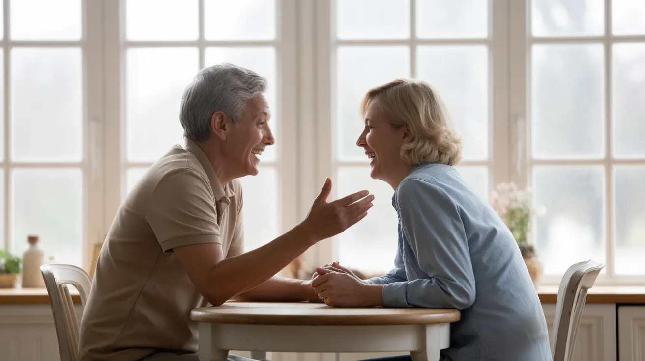 A senior man and woman share a laugh while sitting at their kitchen table, illuminated by soft, natural window light.
