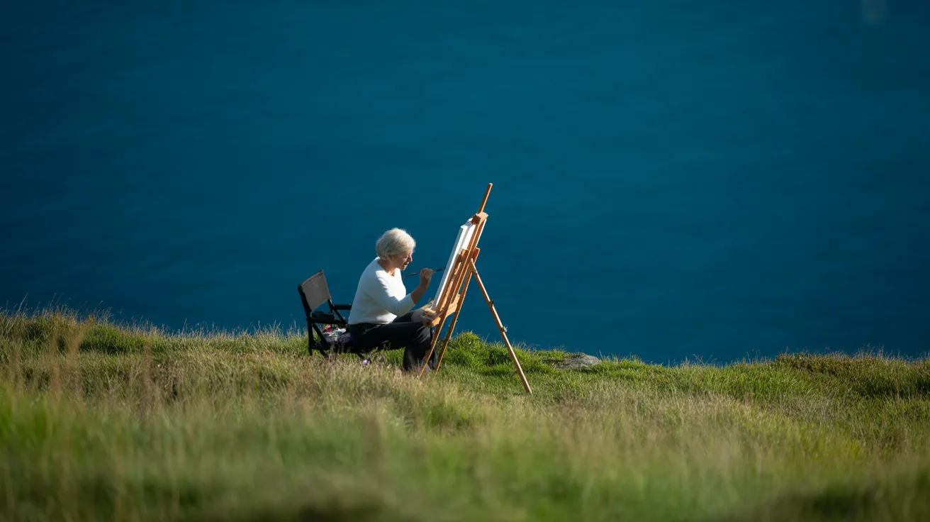 A senior woman paints on a canvas in a vast coastal field under a bright, harsh sun, her shadow long on the grass.