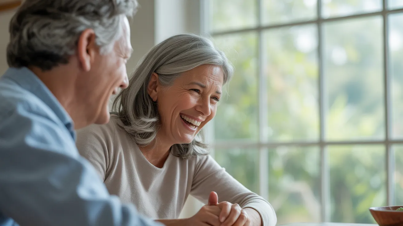 A senior woman in profile laughs warmly while sitting at a kitchen table with her partner, who is seen from behind.