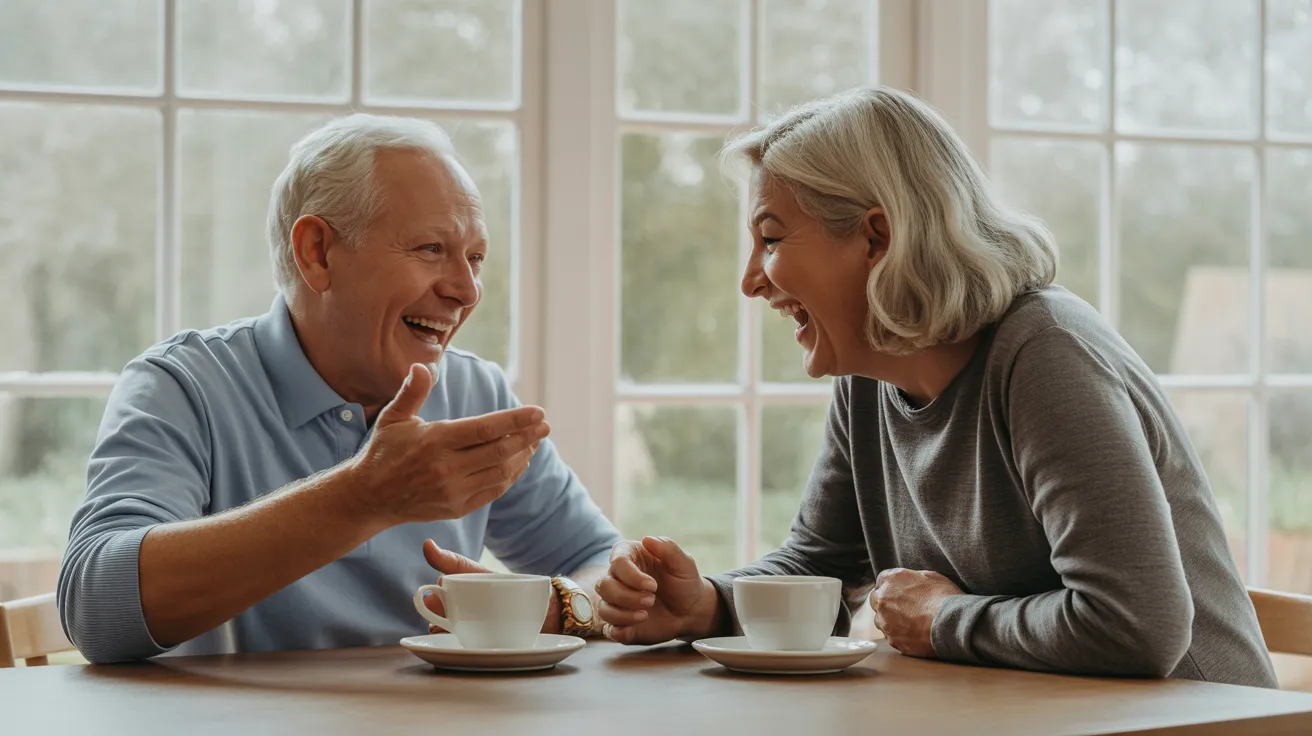 A senior man and woman laugh together while sitting across from each other at a kitchen table, illuminated by soft window light.