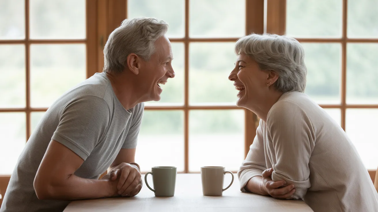 A senior man and woman sit across from each other at a kitchen table, smiling and laughing together over coffee in a brightly lit room.