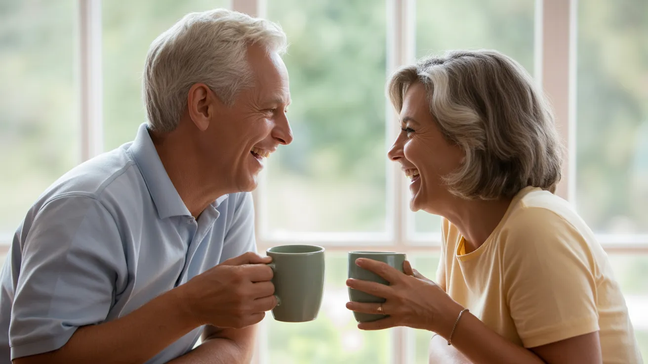 An older couple, a man and a woman, sit at a kitchen table in soft daylight, laughing together while holding coffee mugs.