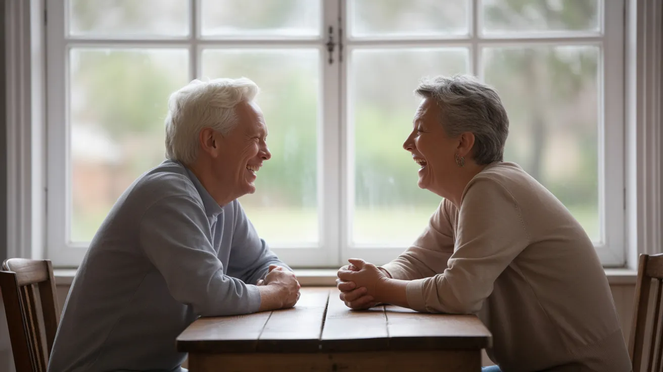 A senior man and woman are laughing together while sitting across from each other at a kitchen table with coffee mugs, lit by soft window light.