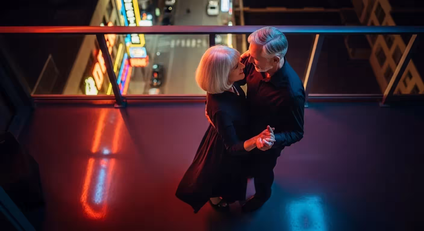 An older couple, seen from directly above, dances closely together on a city balcony at night, illuminated by the warm glow of neon streetlights.
